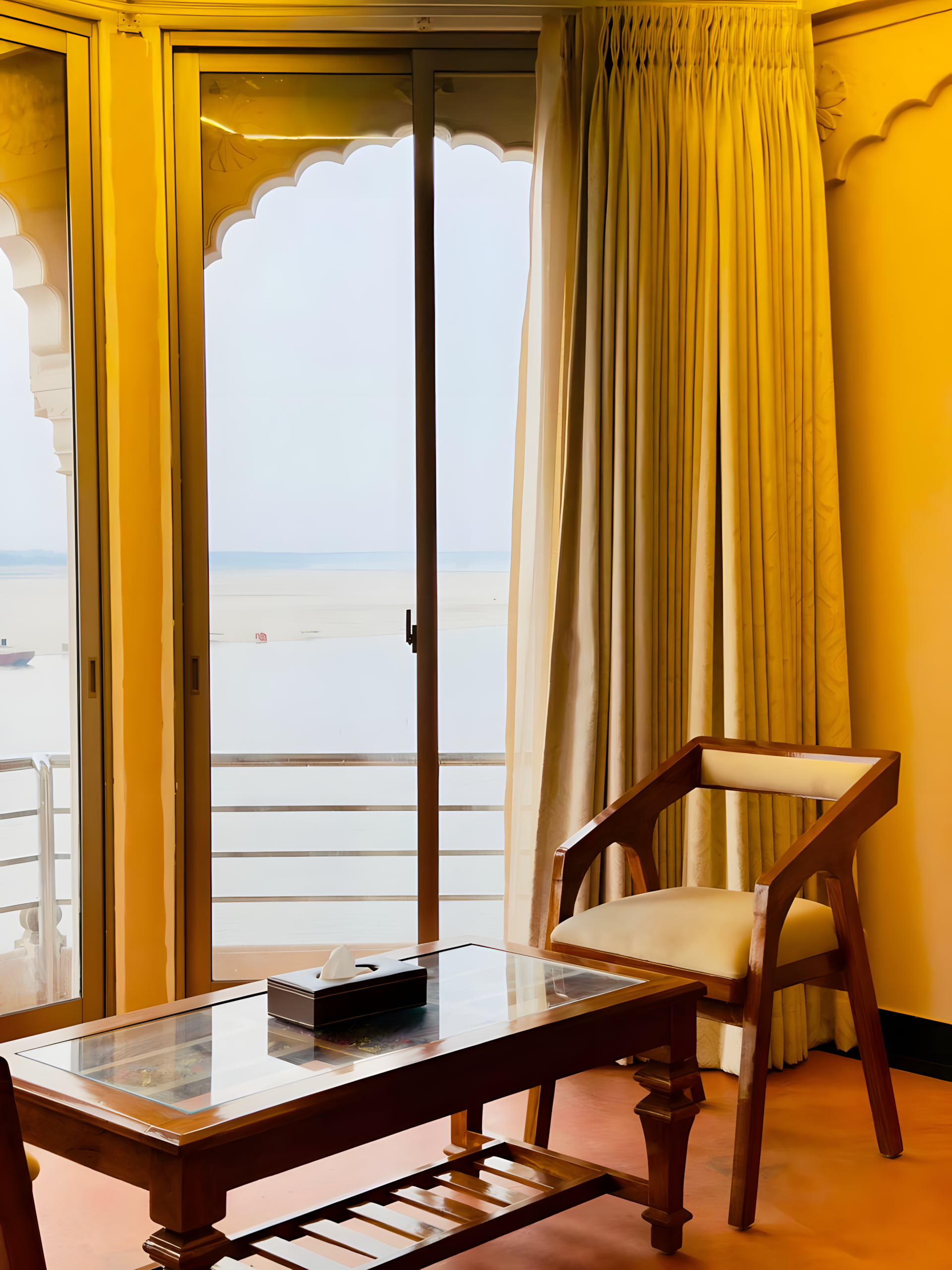 Heritage room interior at Maharana Mahal Varanasi — arched balcony door with yellow ochre walls framing the sacred Ganga river, teak desk and chair — Ganga view room in boutique palace hotel Banaras