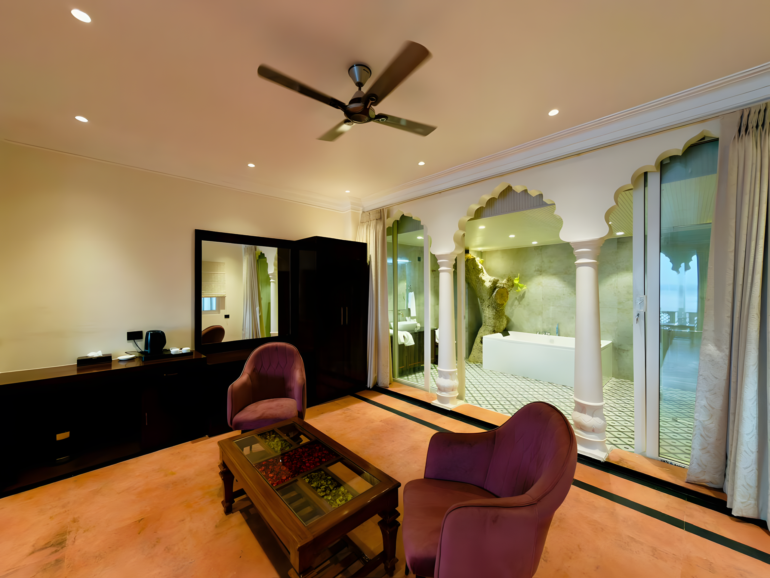 Suite sitting room at Maharana Mahal luxury hotel Varanasi — purple velvet chairs, teak coffee table, open arch view through to spa bathroom with tree-feature soaking tub — boutique heritage hotel Banaras
