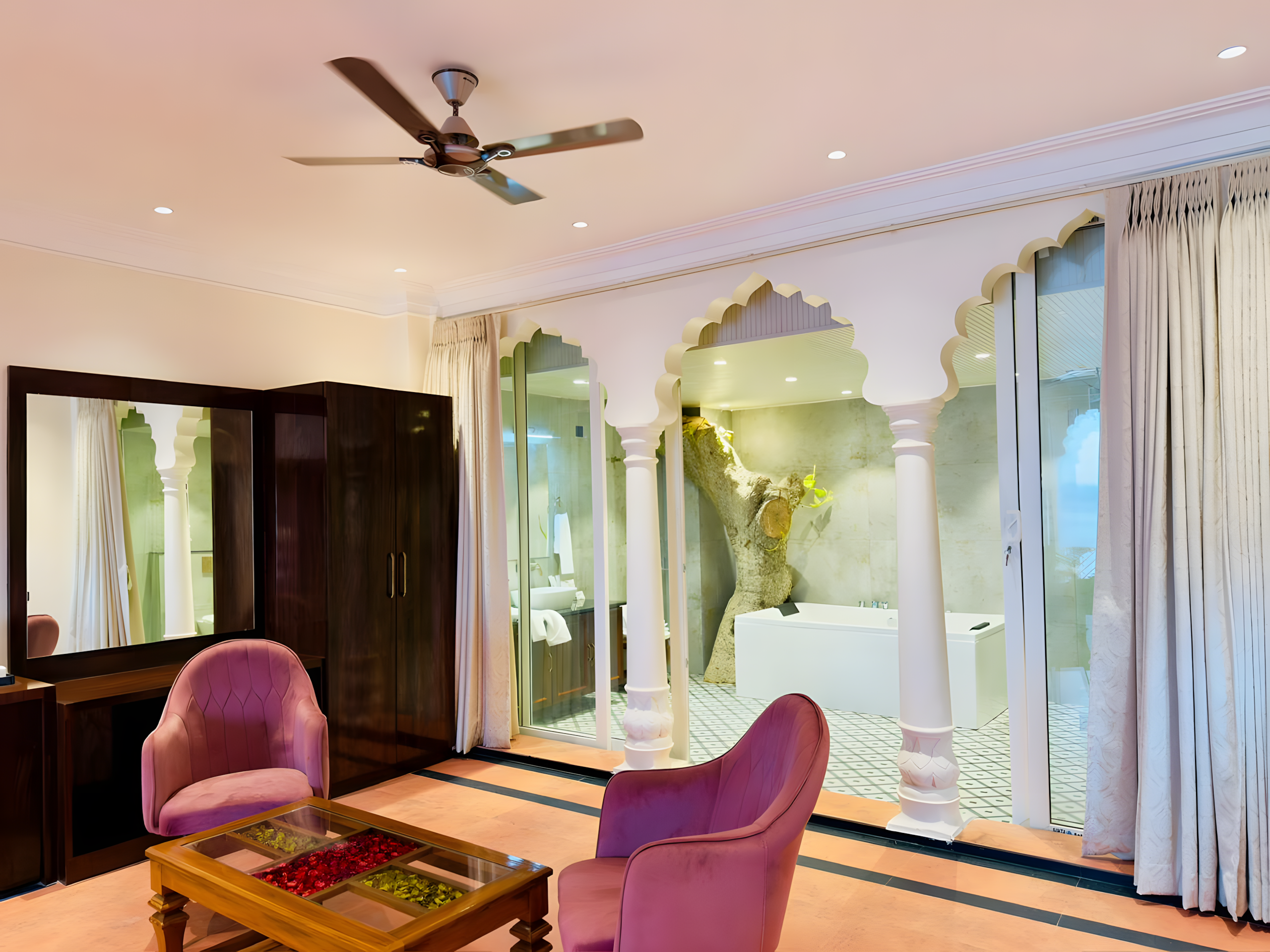 Suite at Maharana Mahal boutique hotel Varanasi — purple velvet armchairs, teak coffee table, open carved arch with glass view into spa bathroom featuring live tree-trunk and soaking tub